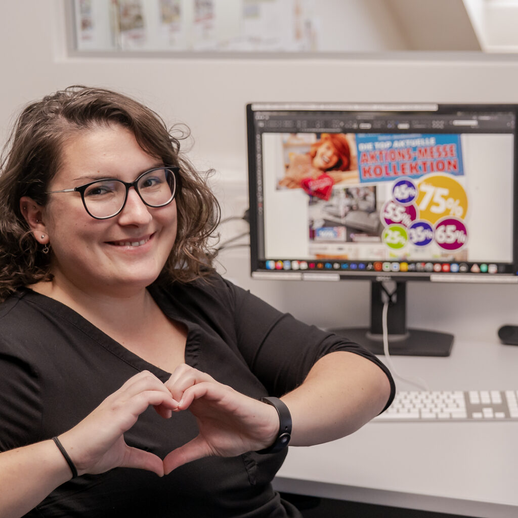 Mitarbeiterin im Büro von Polstermöbel Fischer sitzt vor einem Computer mit geöffneter Grafiksoftware, zeigt lächelnd ein Herz mit den Händen – auf dem Bildschirm eine bunte Werbegrafik mit Prozentzeichen und Aktionshinweis.